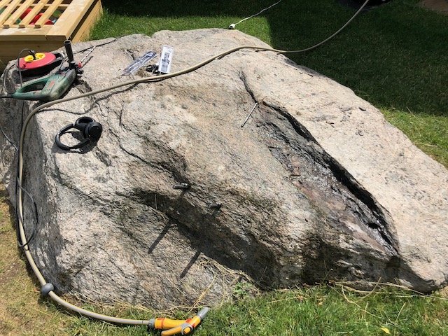 Stor sten med borrade hål och verktyg som kil och hammare för borttagning på gräsplätt.