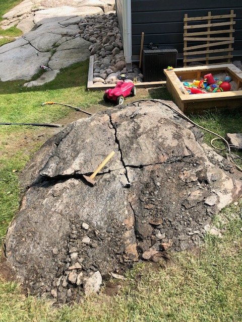Spräckt granitblock i en trädgård med arbetsredskap och leksaker i bakgrunden.