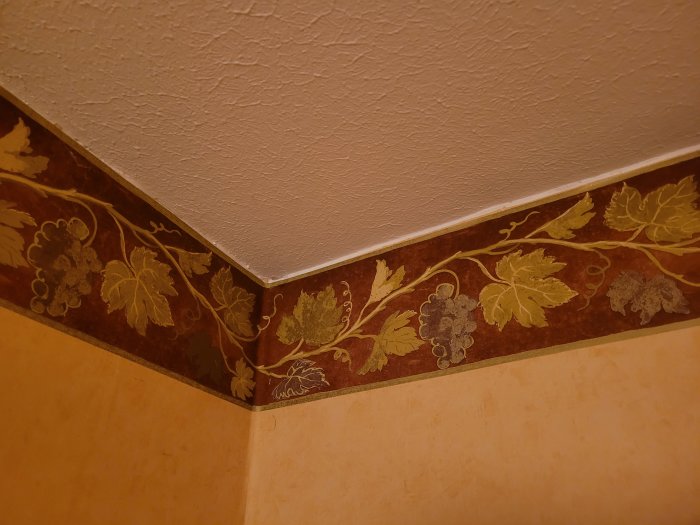 Blommig tapetbård i hörn av rum, äldre tapet under som möter vitmålat tak.