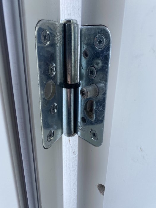 Närbild på öppet metallgångjärn på en vit dörr, skruvar och justeringshål syns.