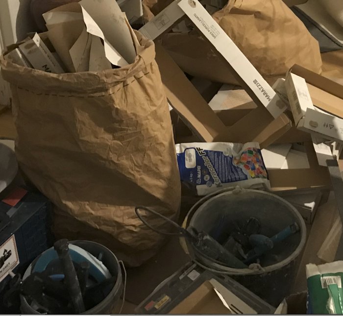Rörigt renoveringsrum med öppna kartonger, byggavfall och verktyg i hink.