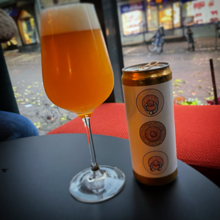 Ett glas fyllt med orangefärgad öl bredvid en burk på ett bord med fönsterutsikt.
