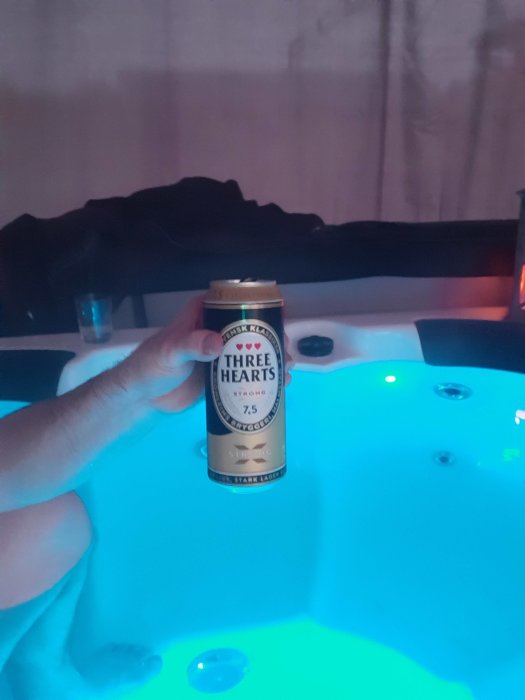 Hand håller en ölburk framför ett bubbelspa med blått ljus och vattenstrålar.