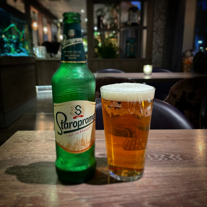 En flaska Staropramen bredvid ett fullt ölglas på ett bord i en bar med suddig bakgrund.