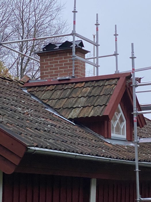 Nyinstallerat väderskydd på tegelskorsten ovanpå rödfärgat hus med tegeltak och byggnadsställning.