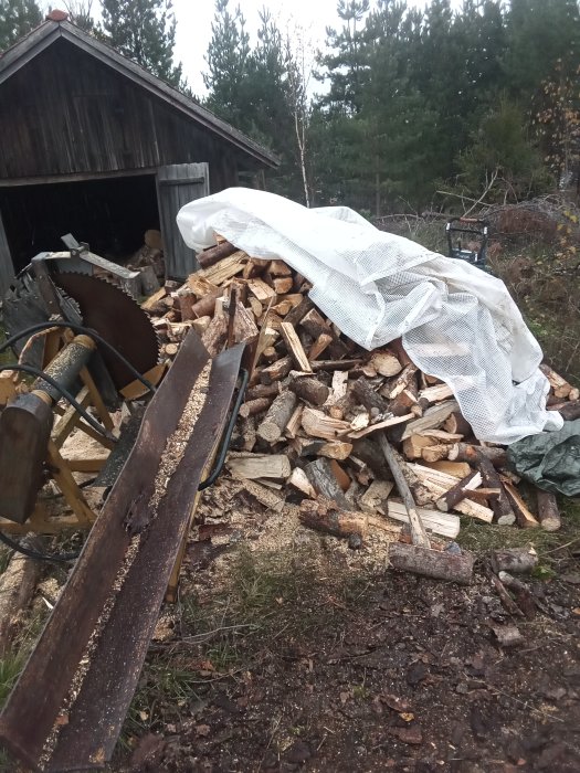 Trave ved delvis täckt med presenning bredvid en vagn och sågklinga, i lantlig miljö.