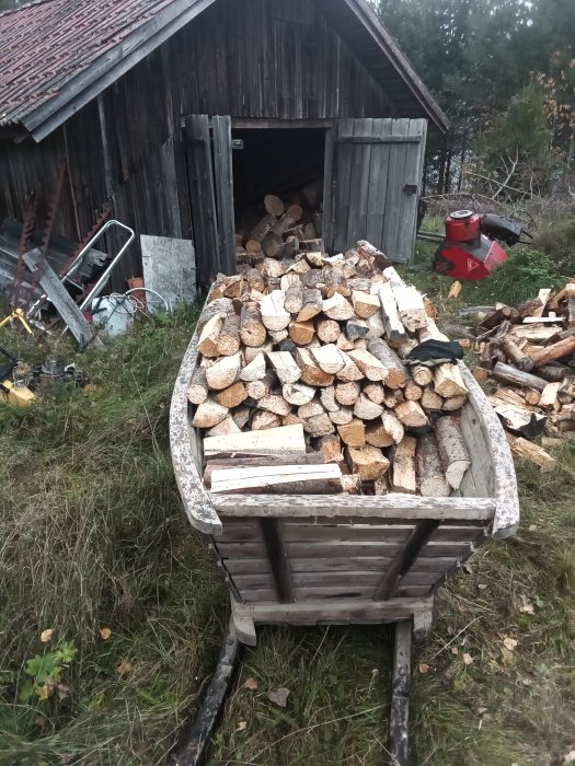 Ved staplad i en gammal släde framför en vedbod, med en röd vedklyv i bakgrunden.