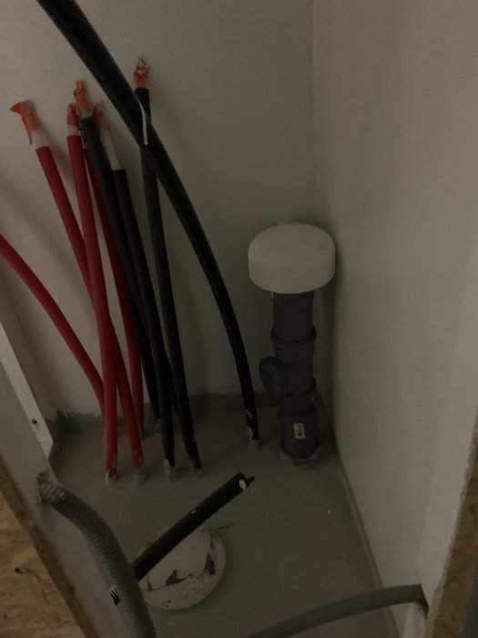 Hörn i ett utrymme med grå och röda PEX-rör, avloppsrör, och en värmekabel, redo för installation.