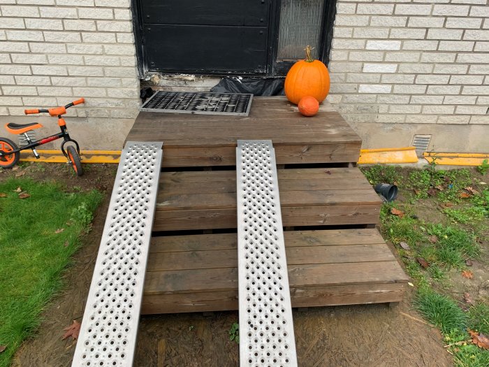 Tillfällig trätrappa utanför hus med metallmattor, pumpa dekoration och barnets cykel i bakgrunden.