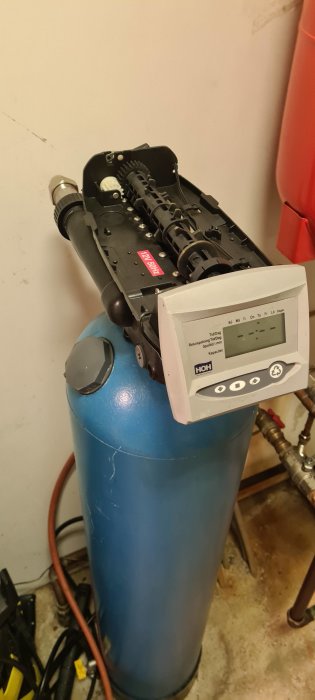 Vattenfilter med digital kontrollpanel och synlig mekanik som ventiler och kamaxel på toppen av en blå behållare.