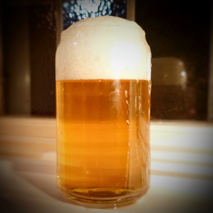 Ett glas fullt med ljus öl och skum på toppen mot en suddig bakgrund.