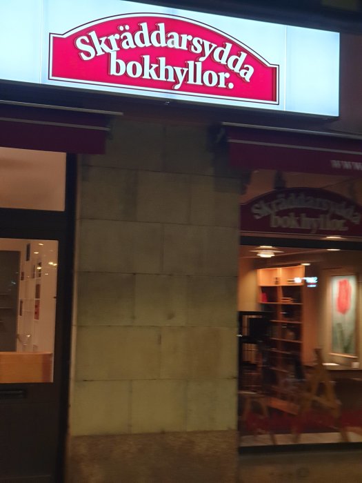 Skylt med texten "Skräddarsydda bokhyllor" ovanför en butiksfönster på kvällen.