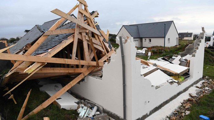 Hus med skadat tak efter storm, synliga takstolar och skiffertak med bråte runt omkring.