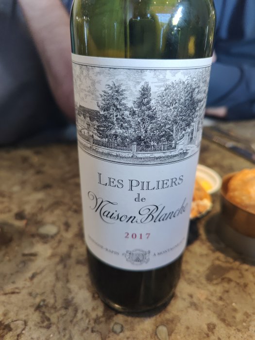 Flaska vin märkt "Les Piliers de Maison Blanche 2017" på ett bord med suddig bakgrund.