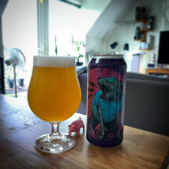 Glas med öl och en ölburk med dinosaurie-motiv på ett bord, inomhusmiljö med suddig bakgrund.