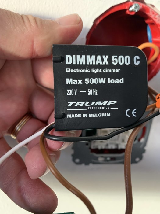 Hand håller en Dimmax 500C elektronisk ljusdimmer med synliga kablar framför en öppen eldosa.