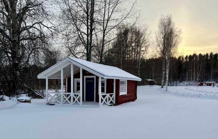 Rödmålat trähus med vita knutar och veranda omgivet av snötäckt mark och träd mot en vinterhimmel.