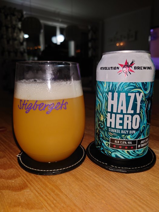 Ölglas med Stigbergets text och burk av Hazy Hero IPA på ett bord, bakgrund av en suddig inomhusmiljö.