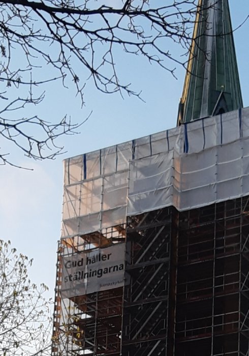 Renovering av kyrktorn med ställningar och skylt som säger "Gud håller ställningarna" mot blå himmel.