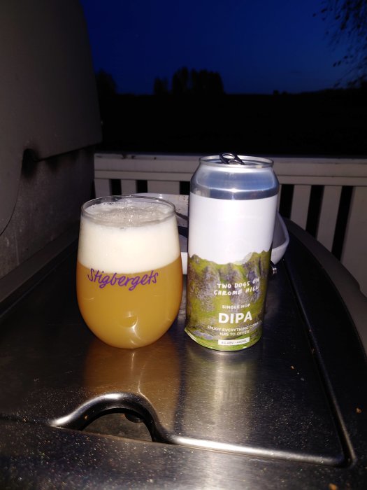 Ölglas fyllt med skummande öl och en ölburk märkt "DIPA" på ett bord utomhus i skymningen.