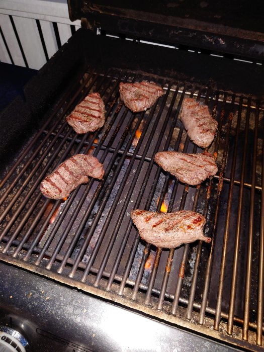 Grillade köttbitar på en BBQ-grill med synlig låga under grillgallret.