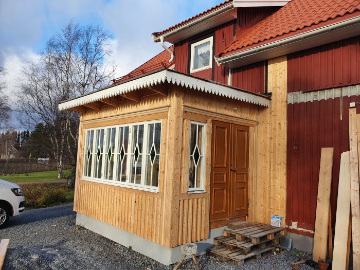 Nybyggd farstukvist med träpanel och snickarglädje, intill rött hus med hängränna.