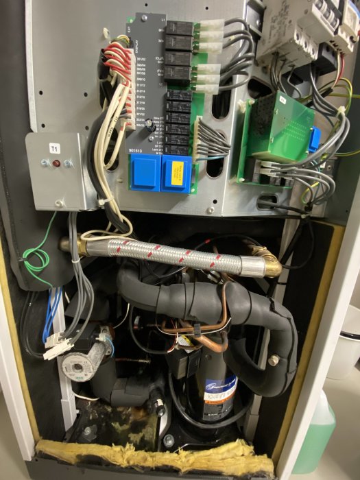 Inre av en värmepump med elektronik, rör, en etikettmärkt knapp T1 och isolering.