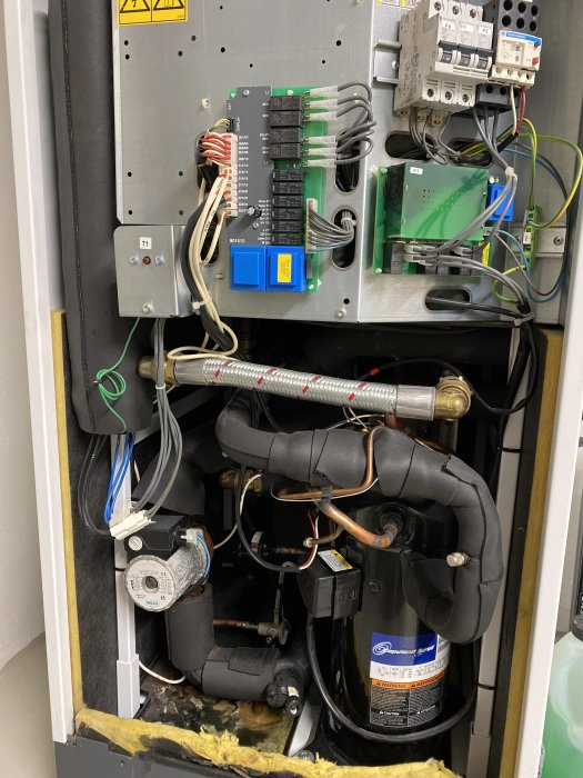Interiör av värmepumpskåp med märkt T1, elkomponenter, pump, ledningar och isolering.