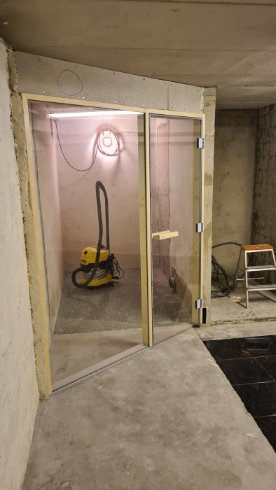 En ouppbyggd ventilation i ett under-renovering utrymme med gipsväggar, en dammsugare på golvet och en svart ring på väggen.