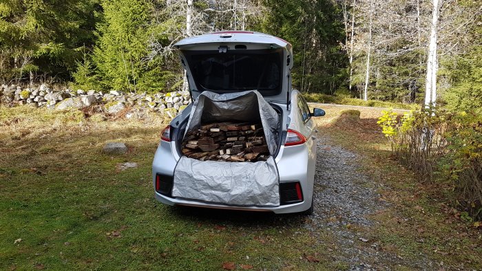 Baksidan av en vit bil öppen med en grå lastsäck fylld med brädor, parkerad på en grusväg omgiven av natur.