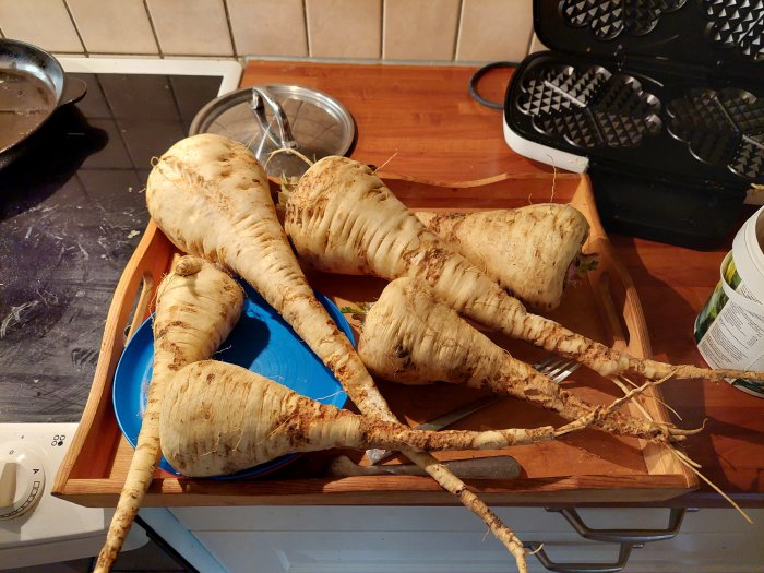 Skördat palsternackor på ett köksbord, bredvid köksredskap och en blå tallrik.
