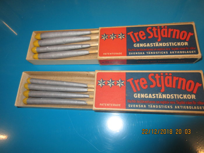Två askar med Tre Stjärnor gengaständstickor på ett blått bord, öppnade för att visa de blå tändstickorna inuti.