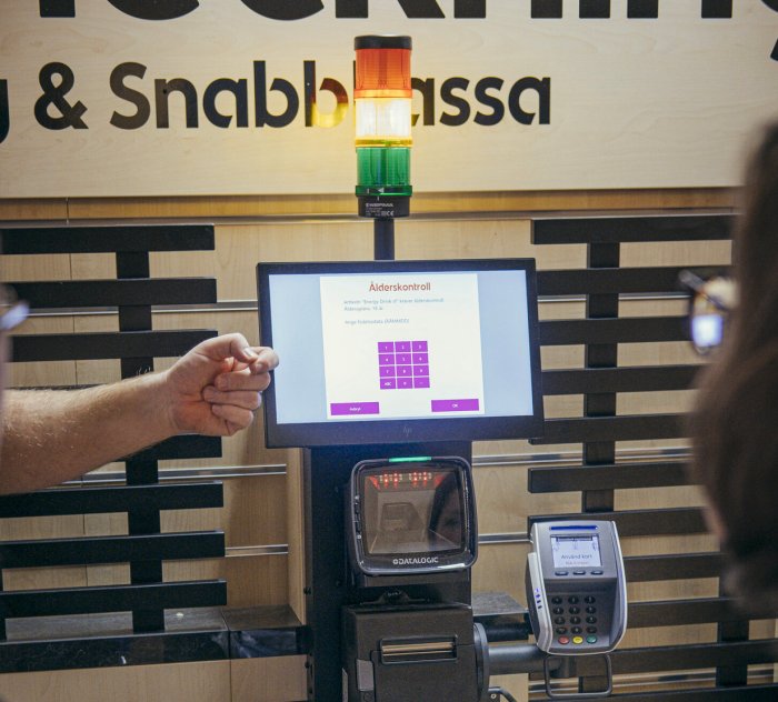 Ålderskontrollsskärm vid självutcheckningskassa med person som pekar, och en signalpelare ovanpå.