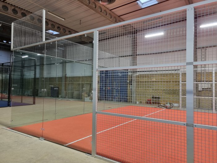 Konstruerad padelbana inomhus med röd konstgräsbeläggning och omgivande glas- och nätstaket.