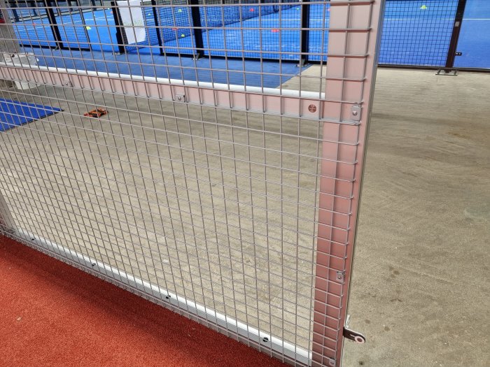 Detaljbild på hörnet av en nyligen konstruerad padelbana med nät, staket och röd markbeläggning.