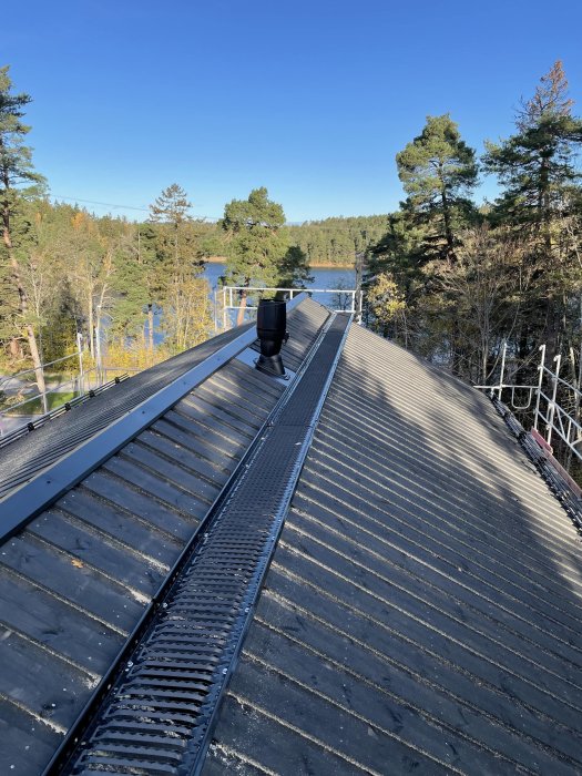 Nyligen färdigställt svart faltak med takintegrerad fläkt, taksäkerhetsräcken och skogssjö i bakgrunden.