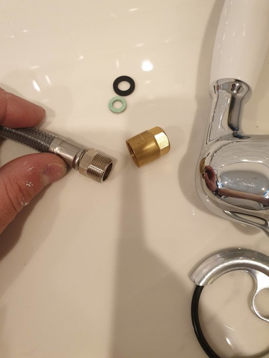 En hand som håller en utdragbar kran, två tätningar och en guld-mutter bredvid en kran på vitt handfat.