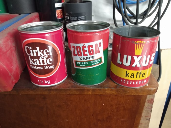 Tre gamla kaffeburkar märkta "Cirkel", "Zoégas" och "Luxus" på ett träbord framför en sladd och röd föremål.