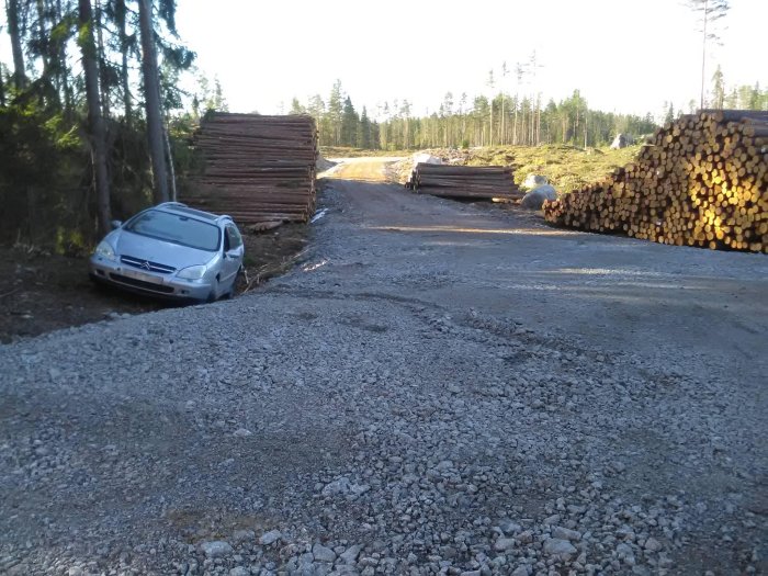 Silverfärgad personbil parkerad olämpligt på grusväg bredvid timmerhögar i skogsmiljö.