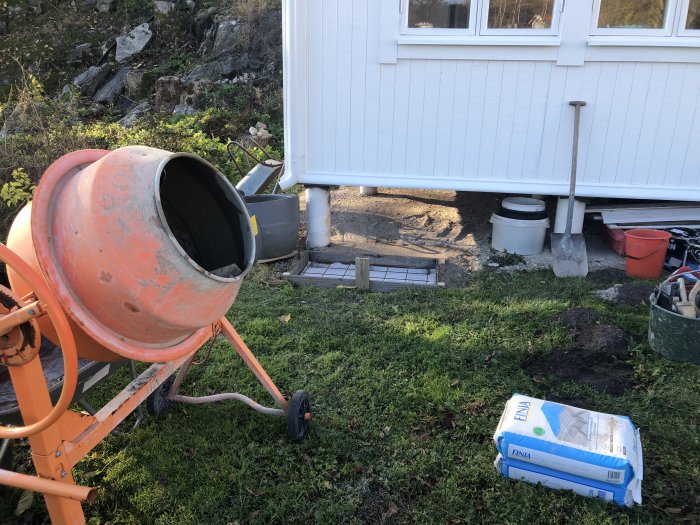 Nyggjuten betongplatta vid husvägg med byggmaterial och en cementblandare i förgrunden.