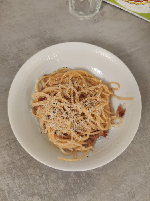 En tallrik med spaghetti carbonara toppad med riven parmesan på ett grått bord.
