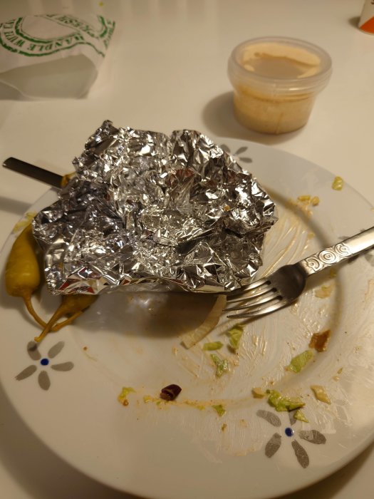 Halväten kebabrulle i folie med en behållare med rosa sås och en fefferoni på en smutsig tallrik.
