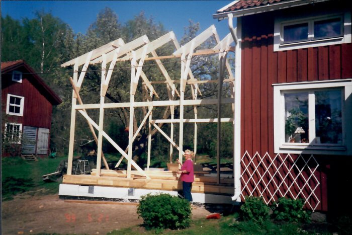 En person står framför en nyuppförd trästomme för en tillbyggnad intill ett rött hus, daterad 1999.