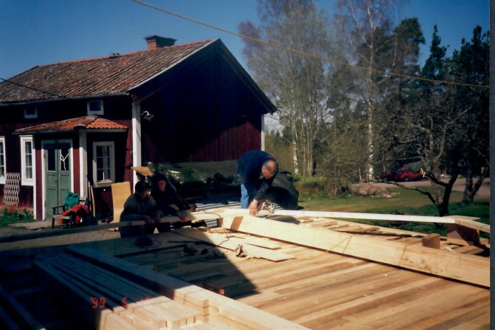 Alt: Personen sågar trä i arbetsprocessen för husbyggnadens stomme utomhus med staplar av virke och hus i bakgrunden.