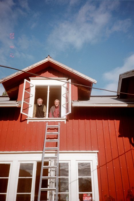 Två personer i öppet fönster på rött hus med taklagskrans ovanför och stege nedanför.