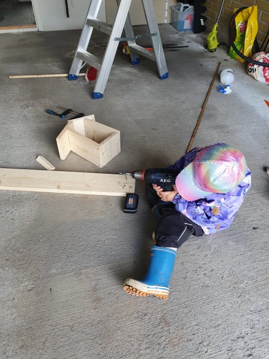 Barn i färgglad mössa använder skruvdragare för att bygga en träfågelholk vid en stege i ett garage.