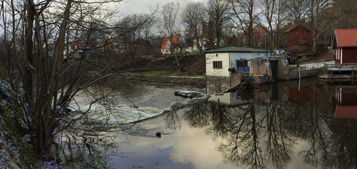 Ett litet vattenkraftverk vid ett vattendrag med omgivande byggnader och spegelblank yta.