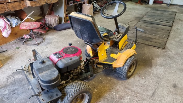 Äldre modell av gul och svart Stiga Park åkgräsklippare i ett garage med motor synlig och sitsen trasig.