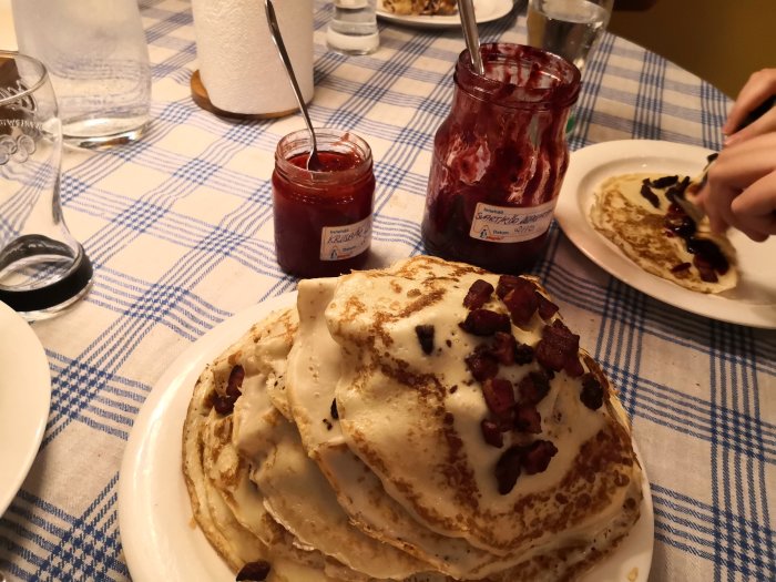 Stapel med tunna pannkakor med knaprigt fläsk, hemgjord sylt och en person som breder sylt på en pannkaka.