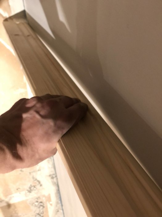 Hand mäter ojämnhet på nyspacklad och reparerad innervägg i betong, med fokus på väggens yta och listverk.